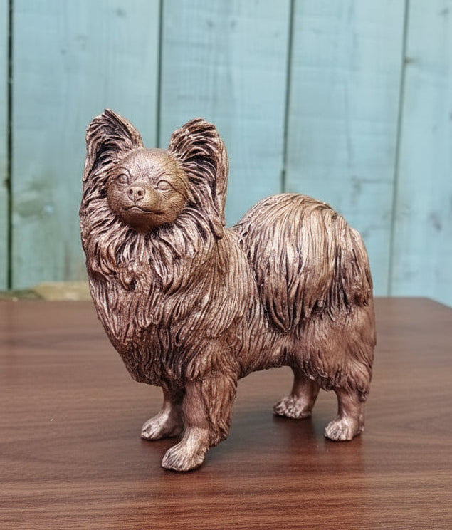 Bronze statue of a Papillon dog on a wooden surface with a light blue wooden panel background